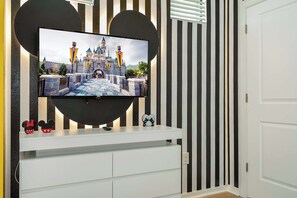 A flat-screen TV mounted on a wall with a Disney castle image, a white entertainment unit below, and black and white striped wallpaper. - Cozy 3BR Apartment Storey Lake Resort Near Disney (Kissimmee)