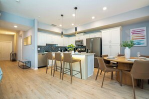 A modern kitchen with white cabinets, a central island with bar stools, a dining area with a wooden table and chairs, and a colorful abstract painting on the wall. - Cozy 3BR Apartment Storey Lake Resort Near Disney (Kissimmee)