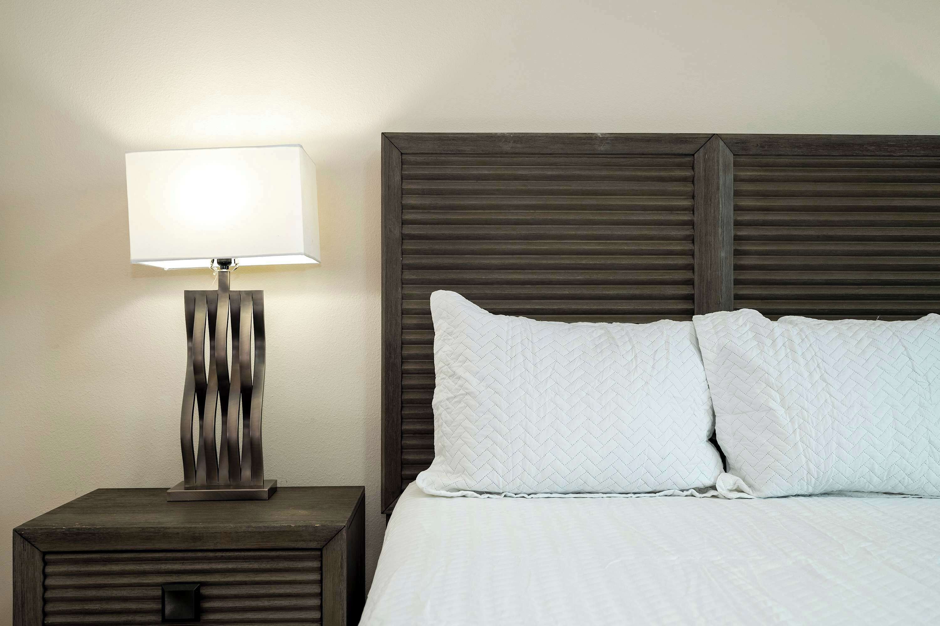 A neatly made bed with white bedding and two pillows, a bedside table with a lamp, and a dark wooden headboard.
