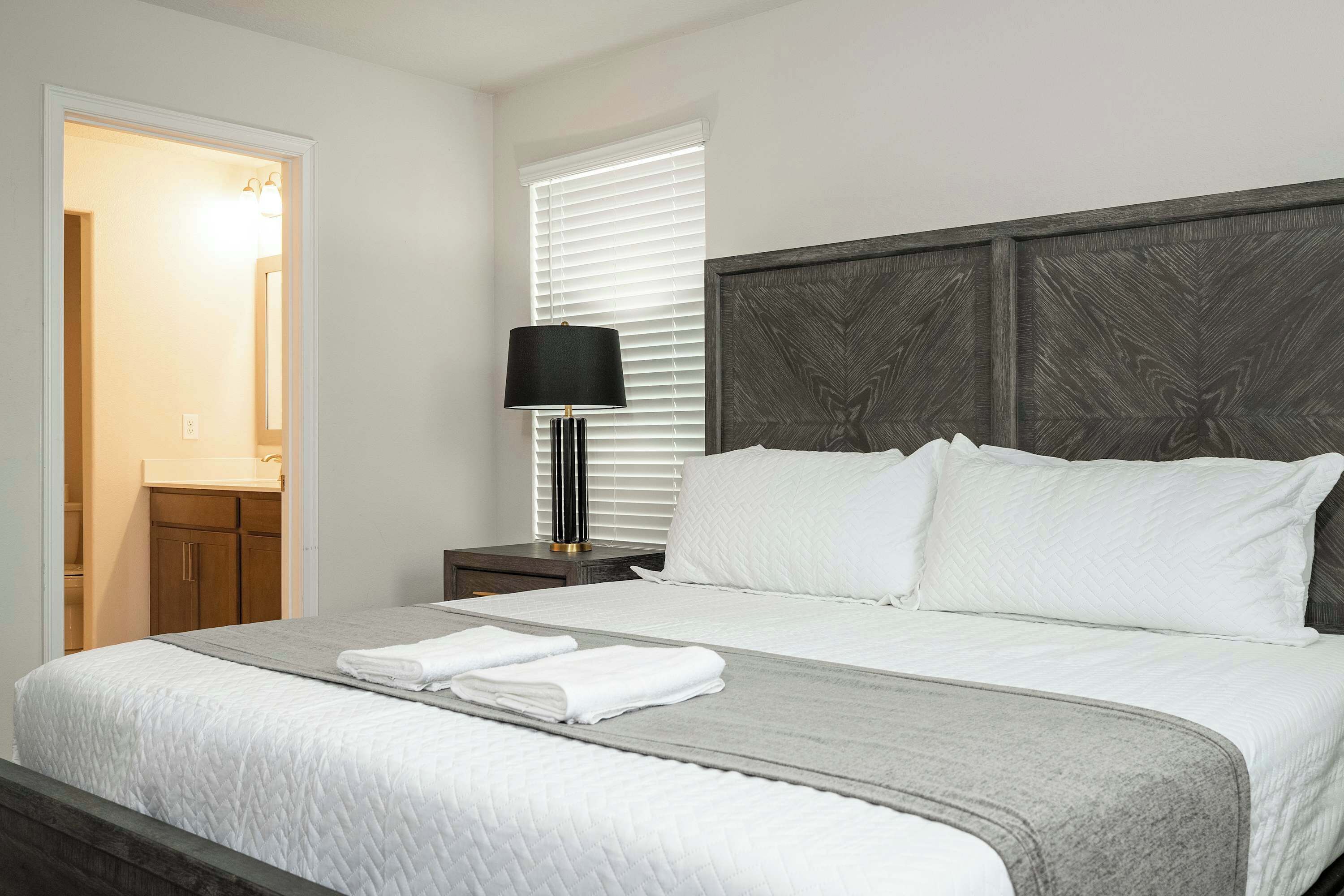 A neatly made bed with white bedding and a grey throw blanket, a bedside lamp, and a dark wooden headboard.