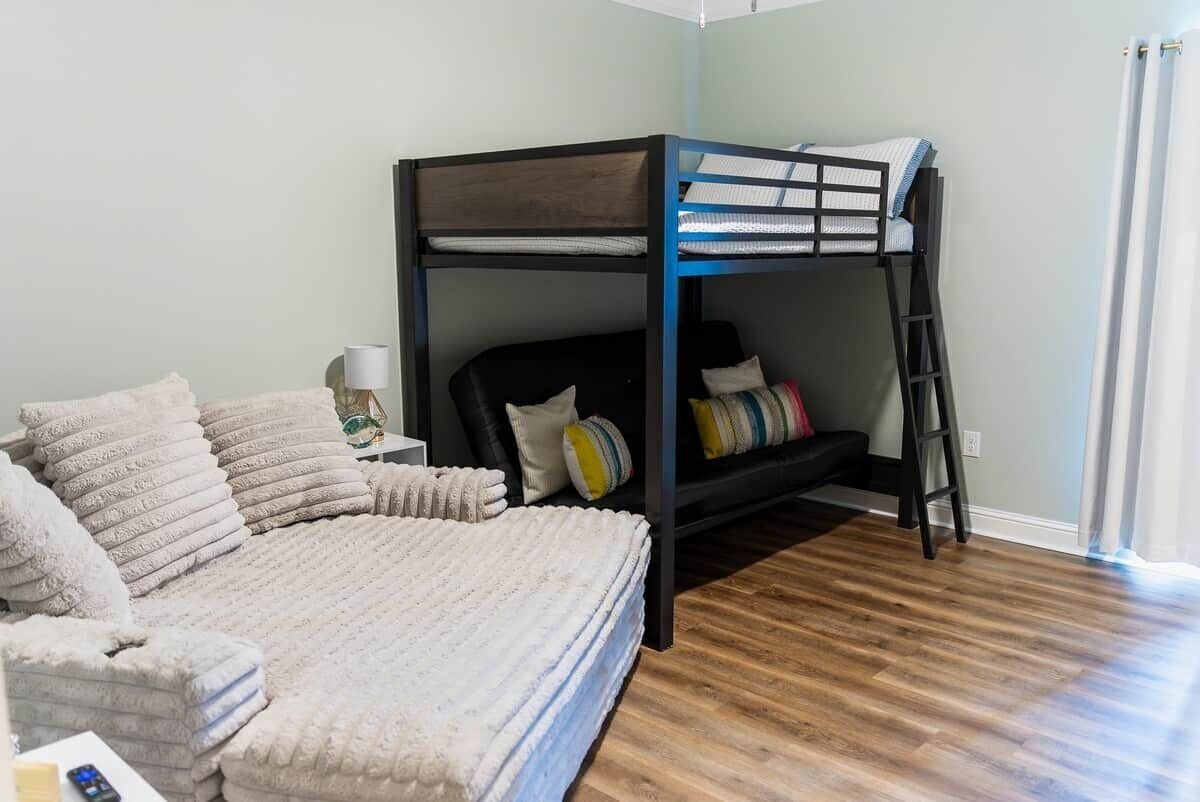 A bunk bed with a ladder, a bedside table with a lamp, and a large bed with multiple pillows.