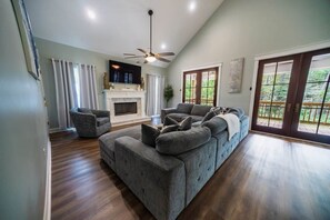 A spacious living room with a fireplace, a large grey sectional sofa, a ceiling fan, and a view through a glass door. - Island Oasis with Included Golf Cart (Dauphin Island)