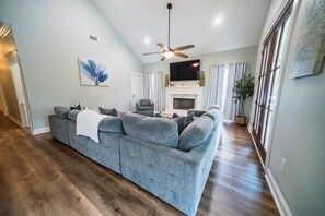 A living room with a large grey sectional sofa, a fireplace, a ceiling fan, and a television mounted on the wall. - Island Oasis with Included Golf Cart (Dauphin Island)