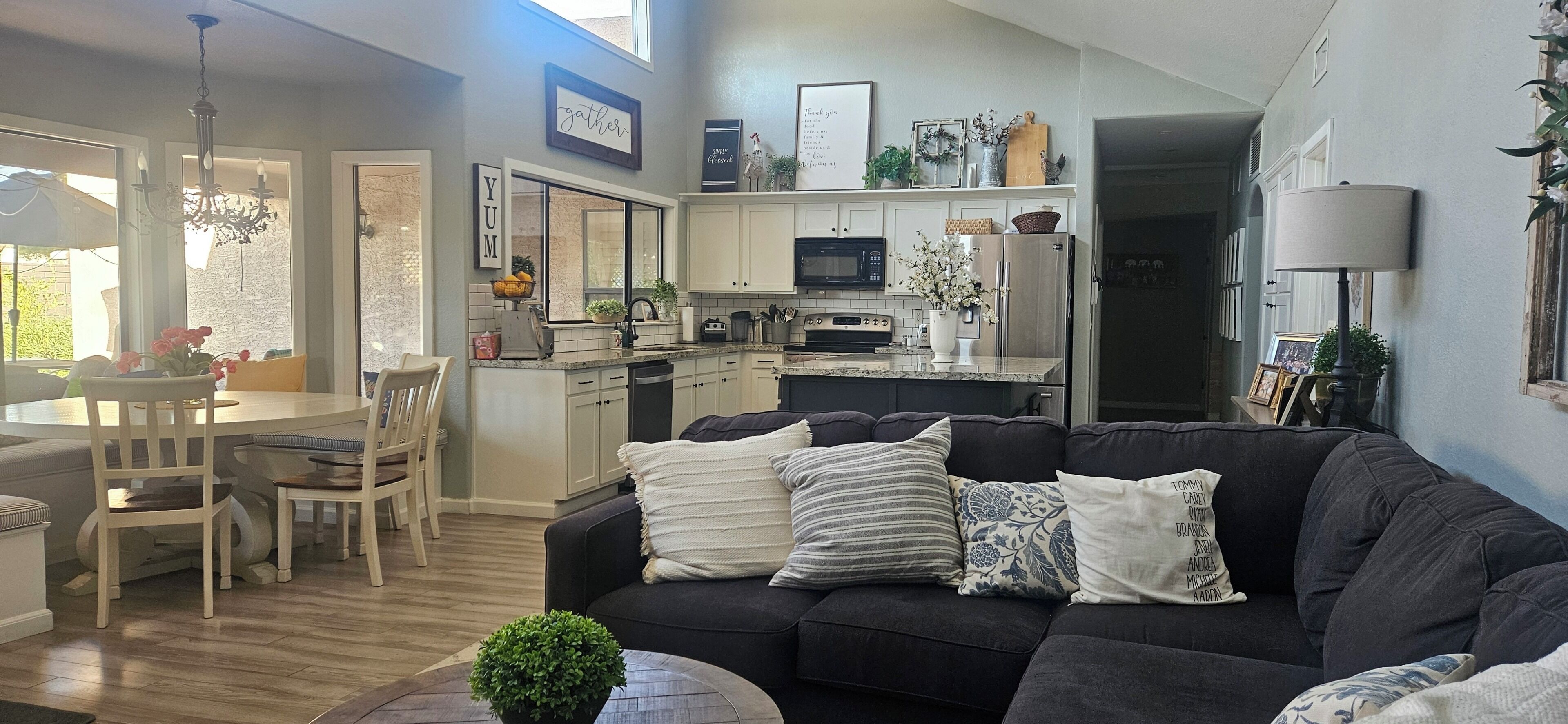 A modern living room with a dark-colored sofa, a dining area with a table and chairs, and a kitchen with white cabinets and a microwave.