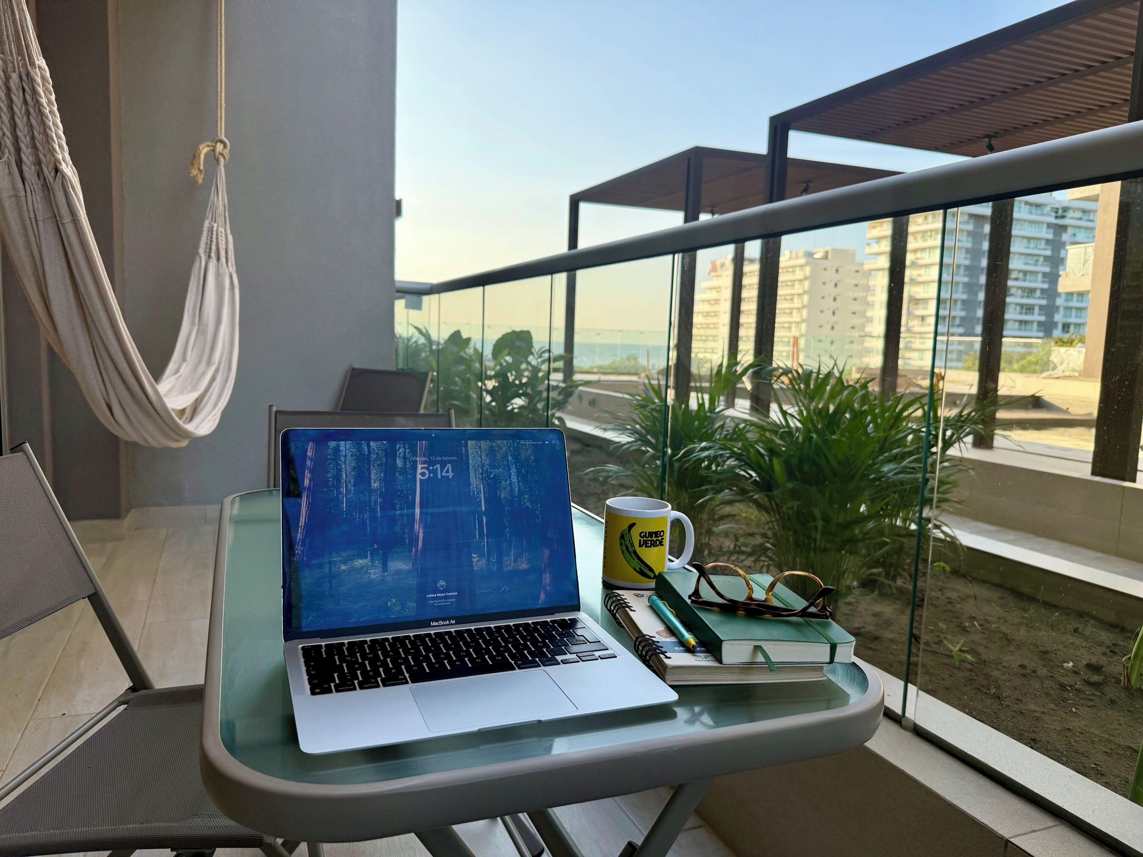 Un MacBook Air sobre una mesa de vidrio, con una hamaca, una taza y un cuaderno en un balcón con vistas a edificios y follaje.