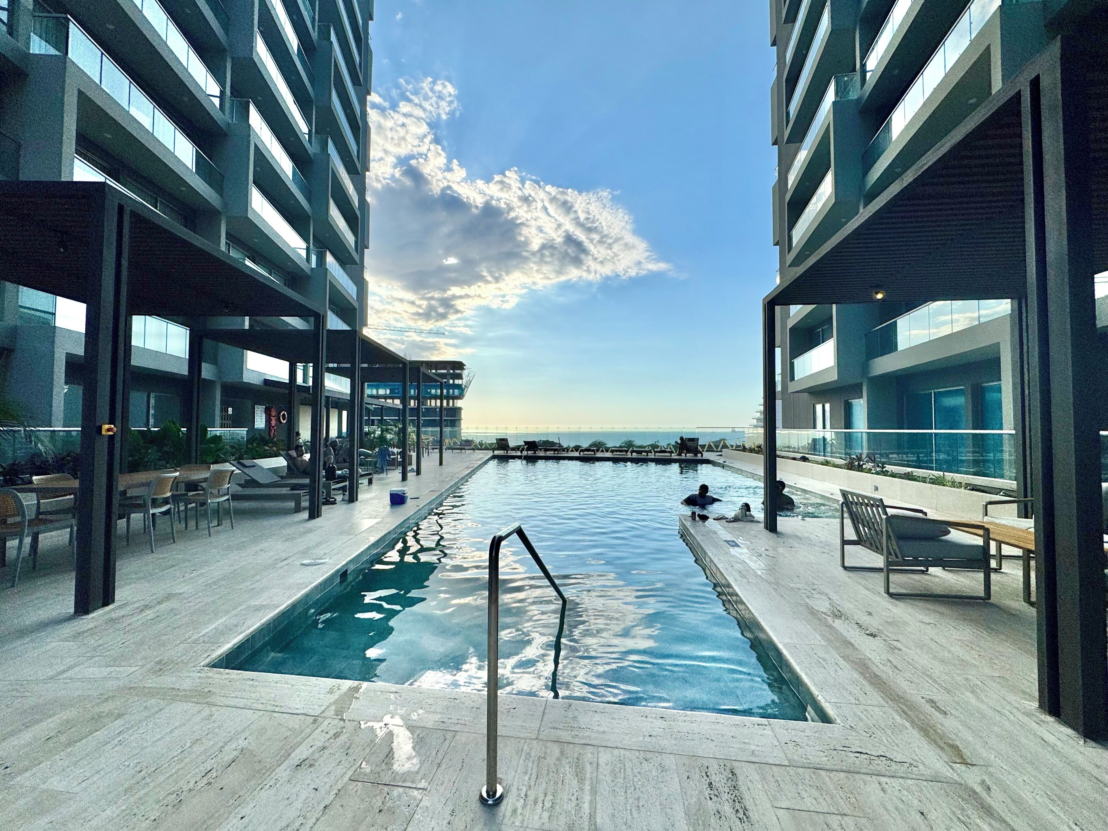 Zona de piscina moderna en un hotel, con vista despejada al mar, sillas de descanso y piscina cubierta.