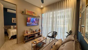 Un salon moderne avec un canapé, une table basse, un téléviseur fixé au mur et une grande fenêtre avec des rideaux.