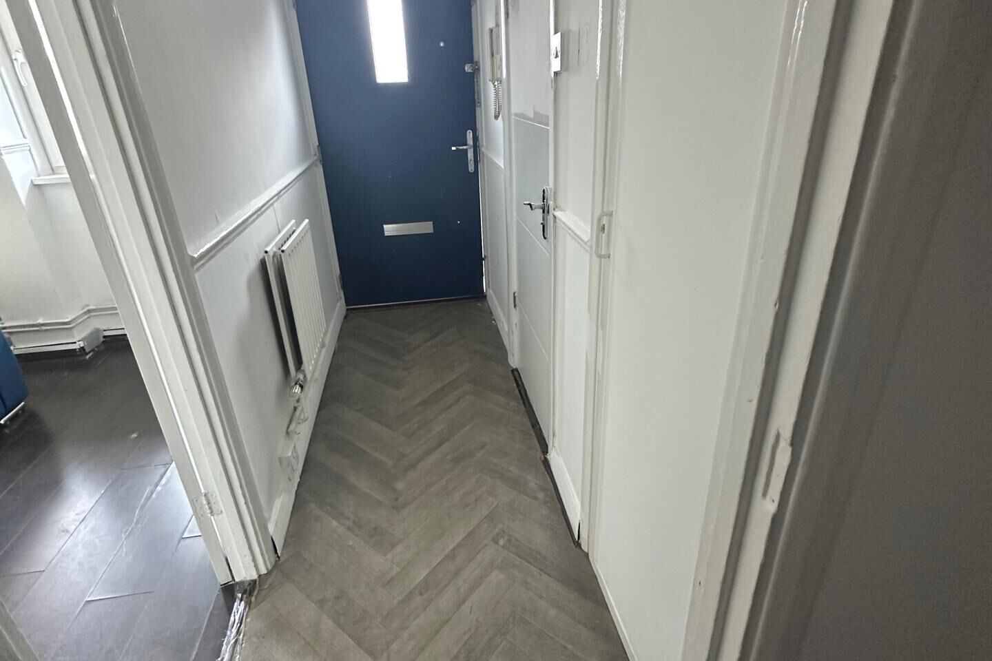 A narrow hallway with a herringbone floor, white walls, and a blue door at the end.