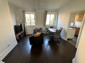 Ett rum med ett mörkt trägolv, en svart soffa med gula kuddar, ett matbord med stolar, en tv och ett köksområde.