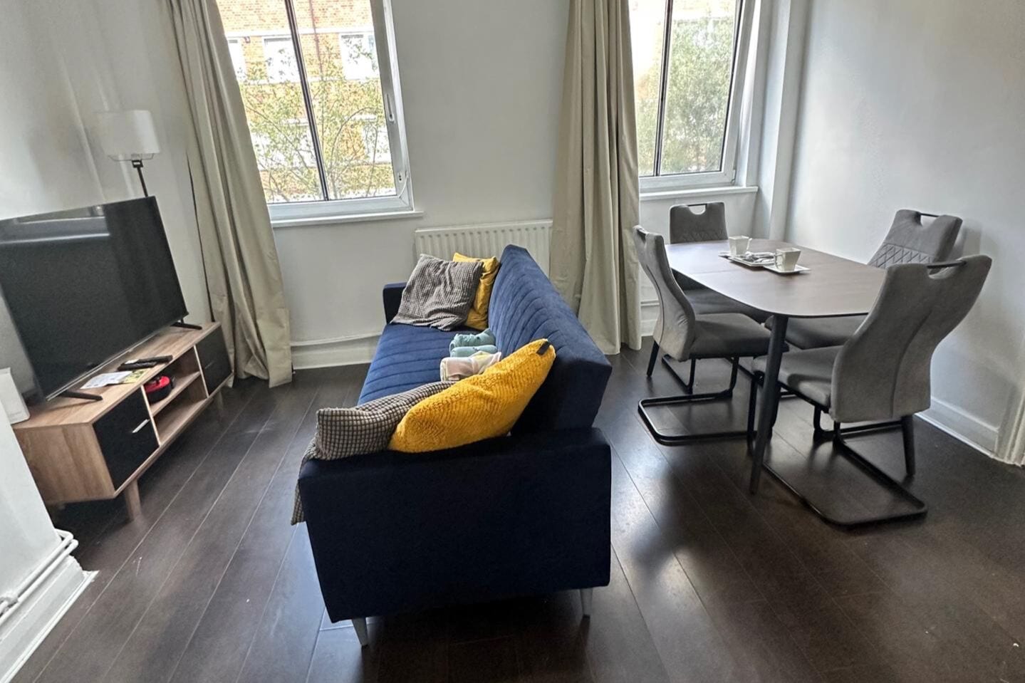 A living room with a dark blue sofa, a television, a dining table with chairs, and a window with curtains.