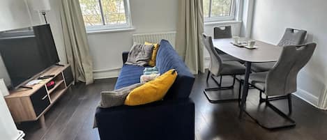 Un salon avec un canapé bleu foncé, un téléviseur, une table à manger avec des chaises et une fenêtre avec des rideaux.