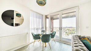 Moderni olohuone, jossa on pyöreä ruokapöytä, vihreät tuolit, harmaa sohva ja suuri ikkuna, josta avautuu näkymä kaupunkiin.
