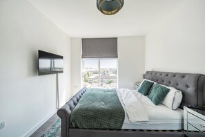 Une chambre à coucher avec un lit, un téléviseur fixé au mur et une fenêtre donnant sur des immeubles.
