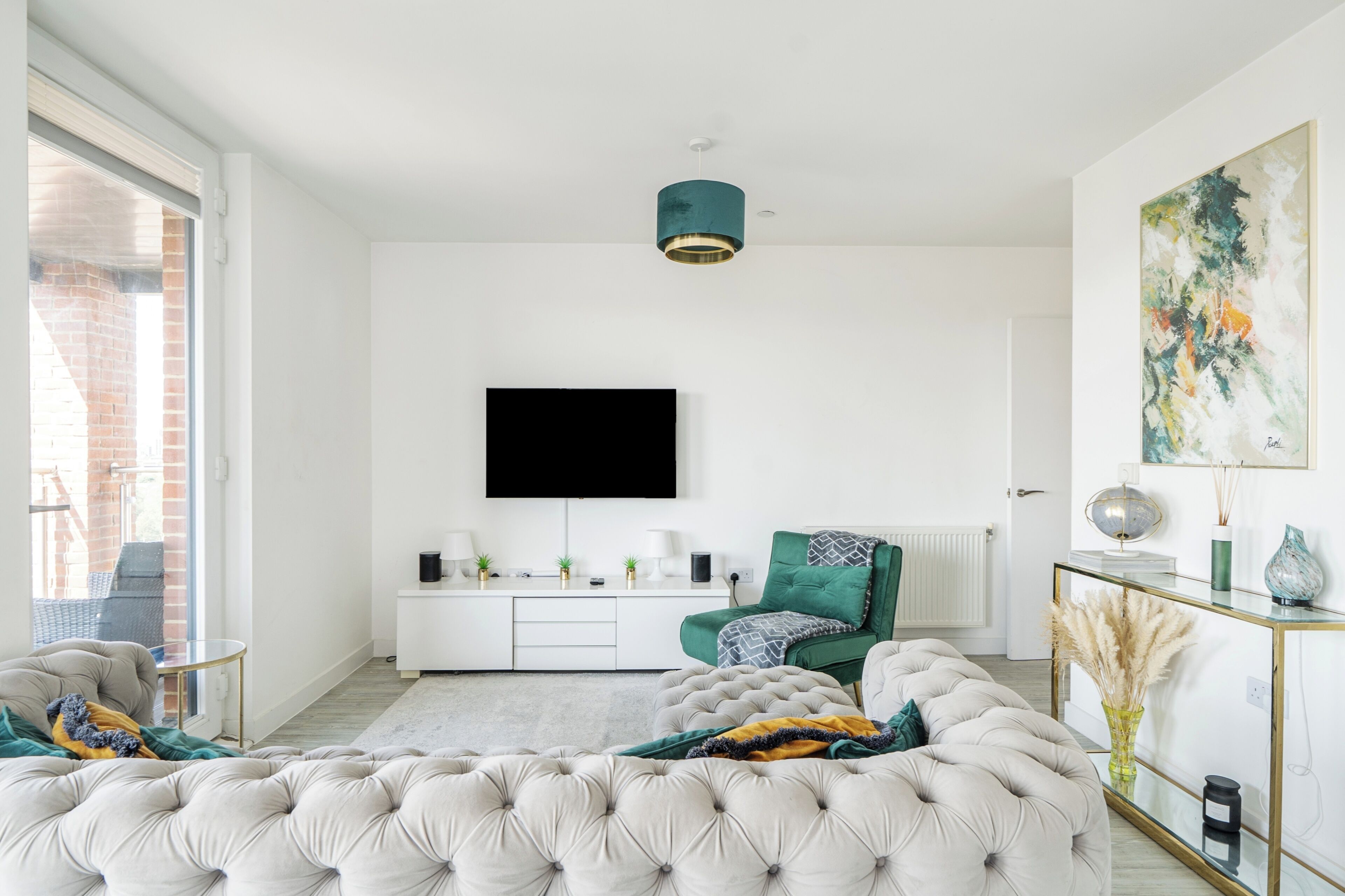 A modern living room with a large tufted sofa, a flat-screen TV mounted on the wall, a green armchair, and a glass coffee table.
