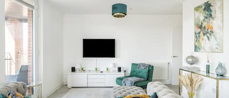 A modern living room with a large tufted sofa, a flat-screen TV mounted on the wall, a green armchair, and a glass coffee table.