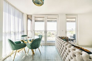 A modern living room with a glass dining table, green chairs, a tufted sofa, and a city view through large sliding doors. - Top Floor Penthouse Near Stadium - Free Parking (London)