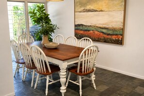 Ein Essbereich mit Holztisch und weißen Stühlen, einer Pflanze in einer Vase und einem großen abstrakten Gemälde an der Wand.