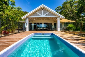 A swimming pool with a wooden deck, a covered patio area, and lush greenery in the background. - Cairns Beachfront Retreat (East Trinity)