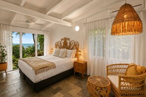 Ein Schlafzimmer mit einem Bett, Nachttischen, einer Hängeleuchte, einem Korbstuhl und Blick ins Freie.