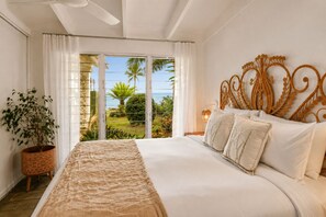 A bedroom with a large bed, a decorative headboard, a potted plant, and a view of a tropical landscape through a sliding door. - Cairns Beachfront Retreat (East Trinity)