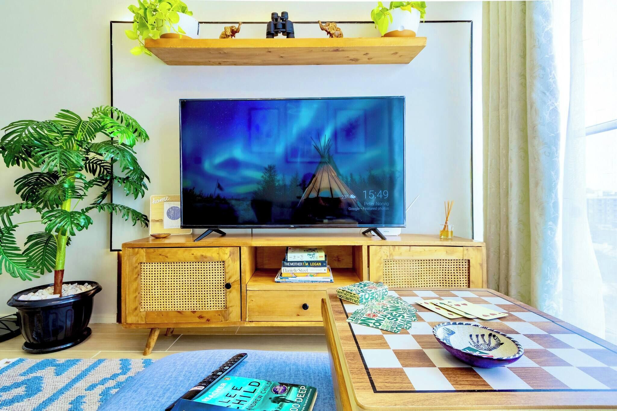 A living room with a television mounted on a wooden cabinet, a potted plant, a small table with a checkered tablecloth, and a window with curtains.