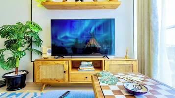 A living room with a television mounted on a wooden cabinet, a potted plant, a small table with a checkered tablecloth, and a window with curtains.
