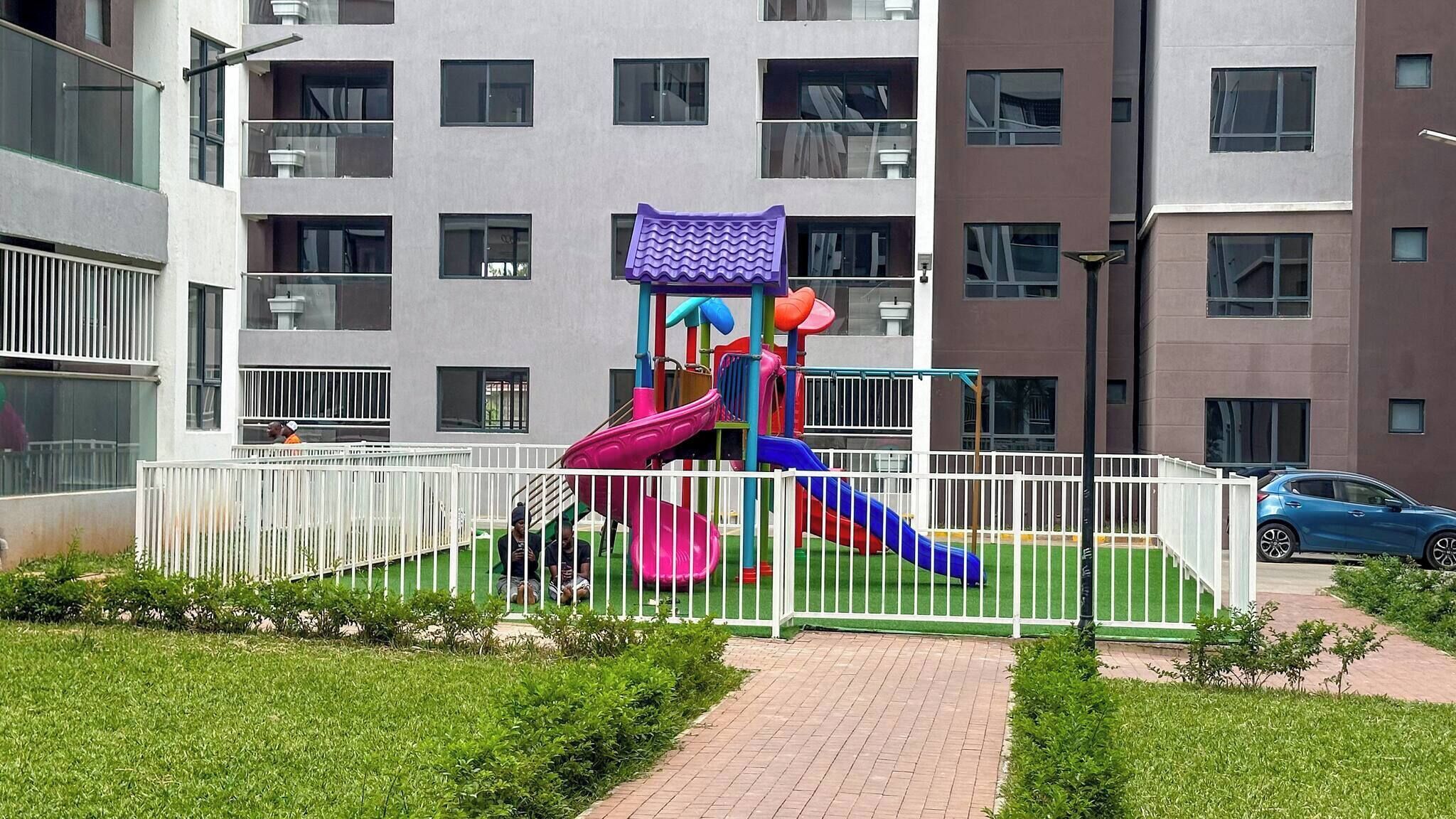 Un parque infantil con toboganes y estructuras para escalar, rodeado por una valla blanca, frente a un edificio de varios pisos con balcones.