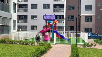 Un parque infantil con toboganes y estructuras para escalar, rodeado por una valla blanca, frente a un edificio de varios pisos con balcones.