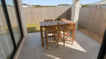 Eine überdachte Terrasse mit Tisch und Stühlen, einem Deckenventilator und Blick auf einen Holenzaun und Grünpflanzen.