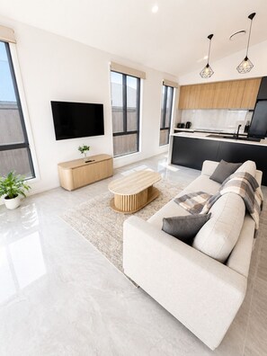 A modern living room with a sofa, a coffee table, a TV, and a kitchen area with wooden cabinets and a black countertop. - Spacious 4 bed 2 bath family home - opposite park - 2 living areas (Greenbank)