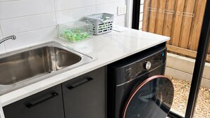 Une cuisine compacte dotée d’un évier en acier inoxydable, d’un lave-linge noir et d’un dosseret de carrelage blanc.