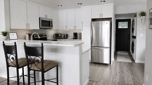 A modern kitchen with white cabinetry, stainless steel appliances, and a central island with two chairs. - `Reflection`s Edge` Lake House & Private Dock! (Port Angeles)