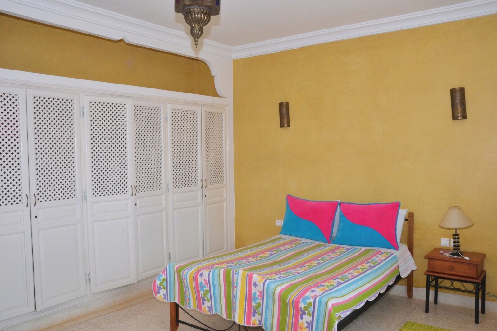 Une chambre à coucher comprenant un lit, une table de chevet, une lampe et une armoire dont la porte est décorative.
