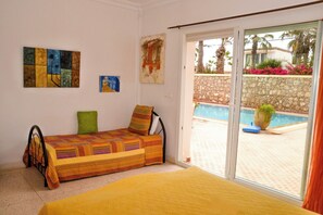 A room with a bed, a sofa, a painting, and a view of a pool and garden. - Vrbo Property (Mirleft)