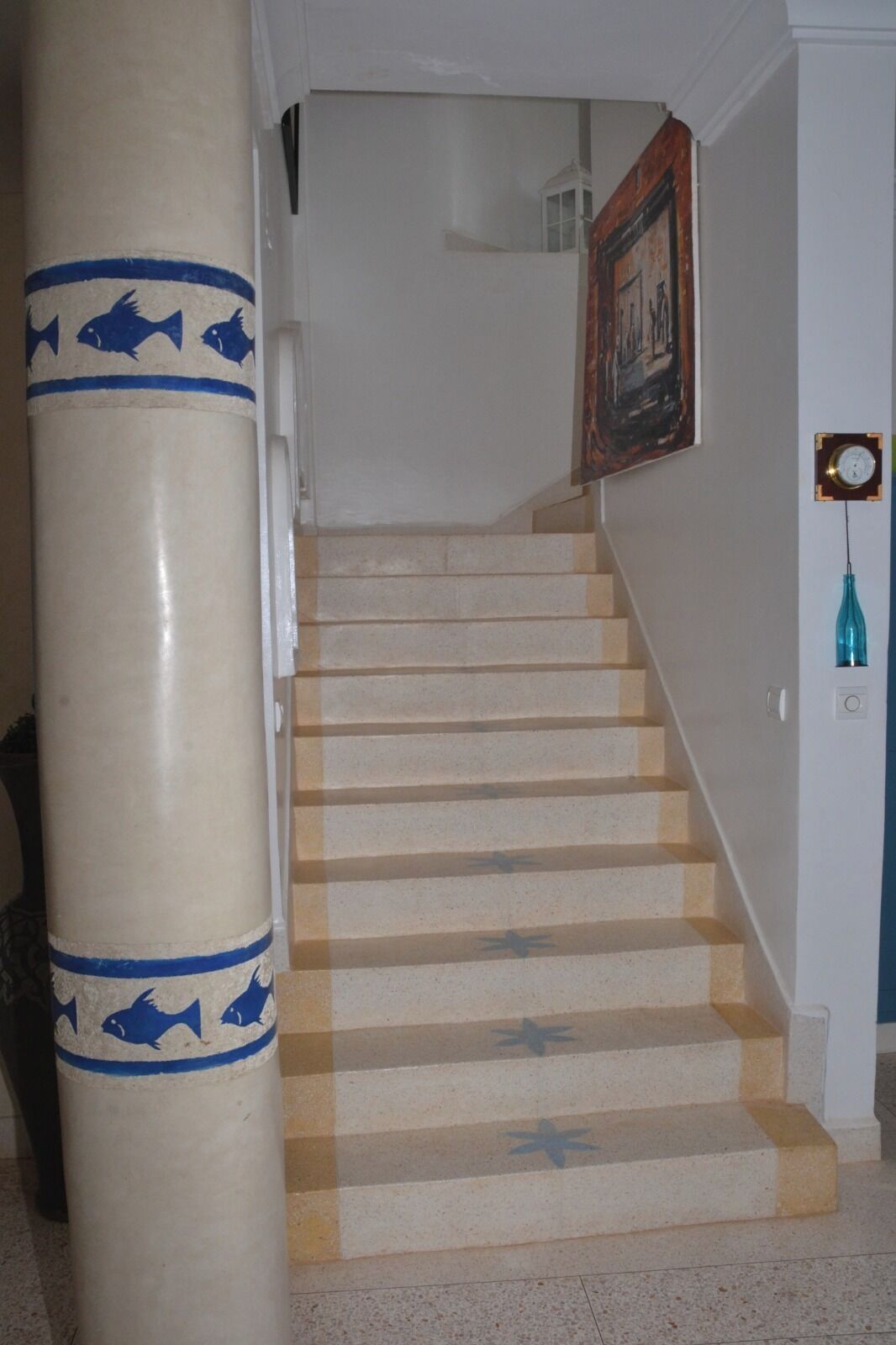 Un escalier aux marches de couleur beige, orné de motifs en forme d’étoiles bleues, menant à un palier où se trouvent une horloge et un tableau encadré accroché au mur.