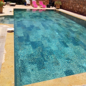 A clear swimming pool with lounge chairs and a stone wall.