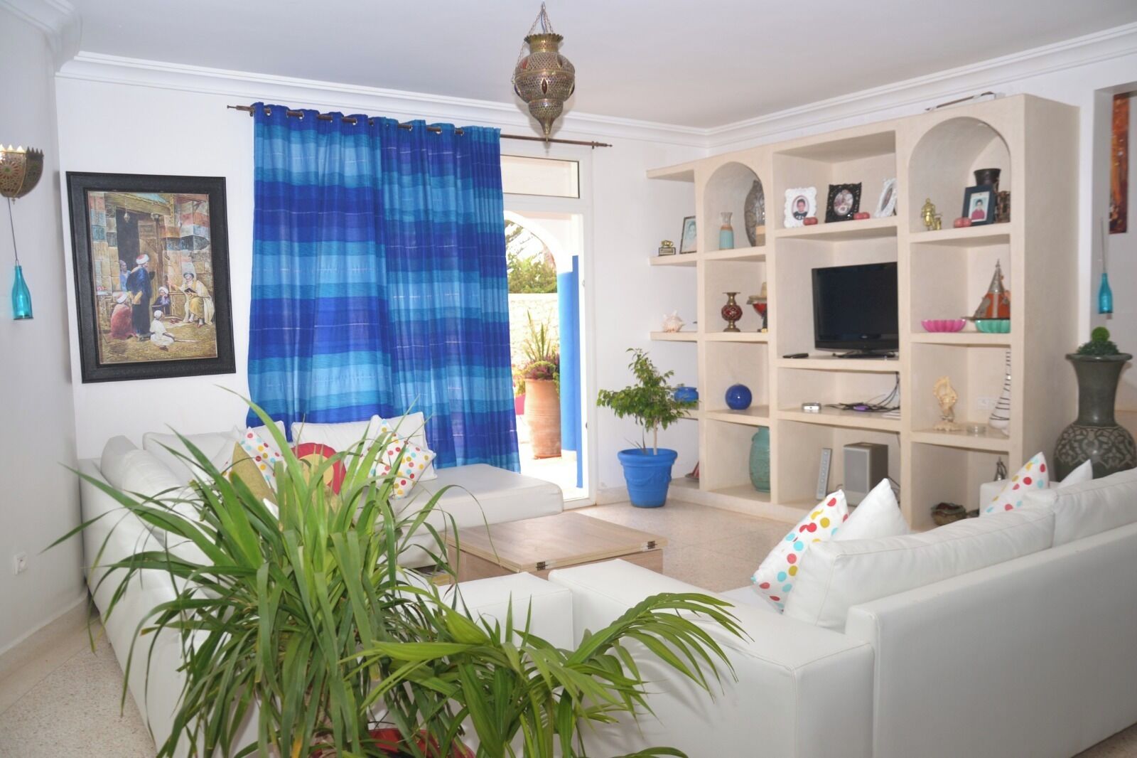 Un salon avec un canapé blanc, un rideau bleu, un téléviseur posé sur une étagère et une plante en pot.