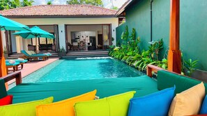 Zona de piscina con sillones de descanso y sombrillas, un edificio con techo de tejas y un muro verde con plantas.