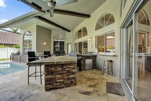 A covered outdoor kitchen area with a stone island, bar stools, and a grill. - Luxury Coral Springs Retreat (Coral Springs)