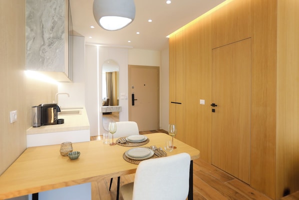 A modern dining area with a wooden table set for two, a kitchen counter with appliances, and a large wooden wardrobe.