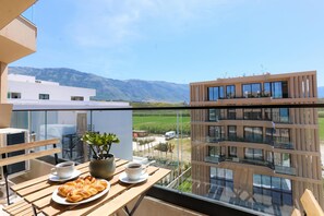 A balcony with a table set for two, a potted plant, and a view of a modern building and mountains. - Modern Pool Side Studio Palm, Vlore, Albania (Vlore)