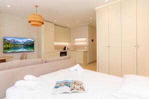 Quarto de hotel moderno com cama, televisão exibindo paisagem de montanhas, cozinha compacta e amplo guarda-roupa.
