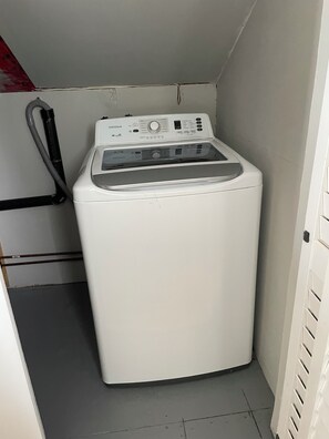A compact, white front-loading washing machine in a small laundry room with a grey floor and a red towel hanging on a hook.