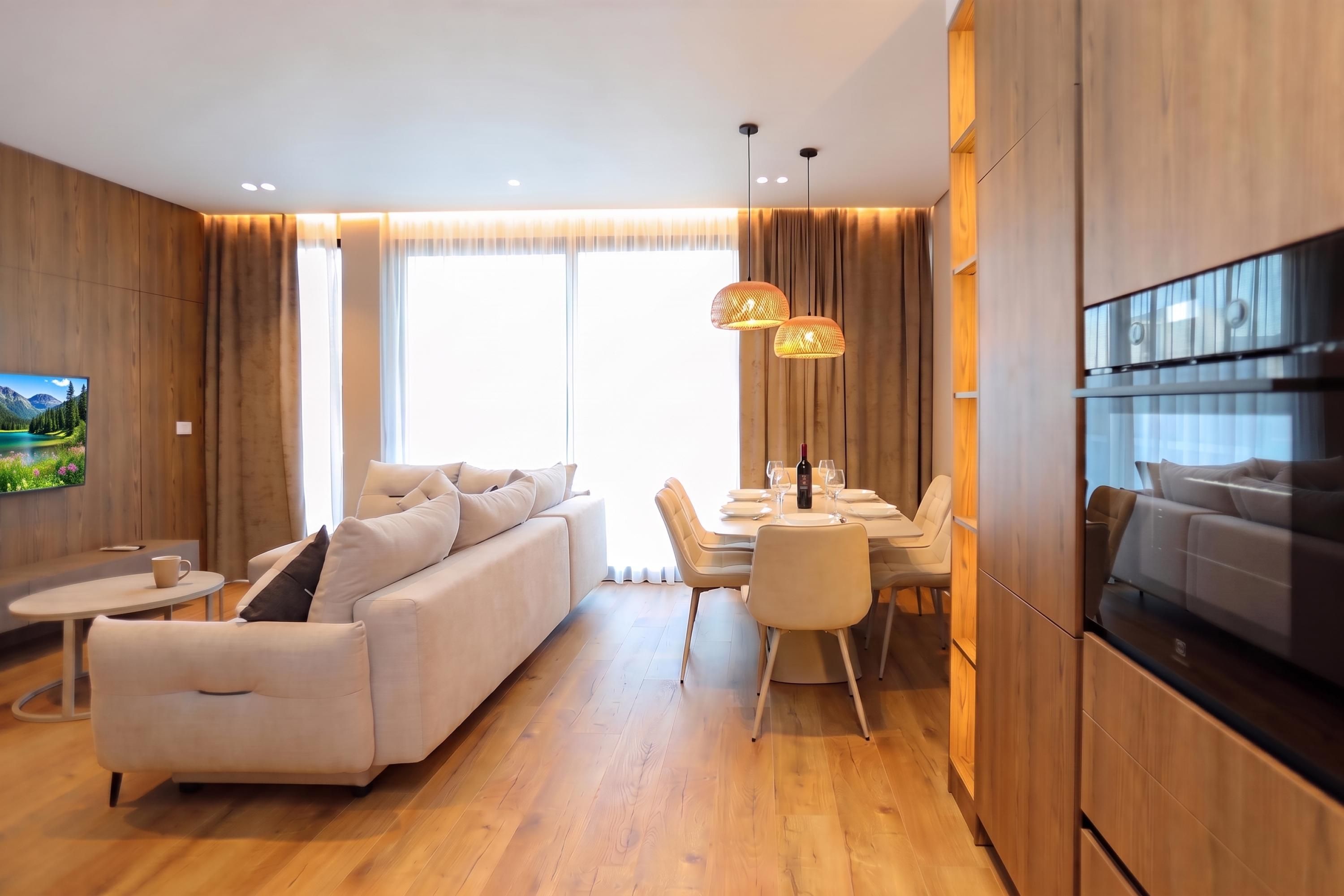 A modern living room with a sofa, dining table, and chairs, a flat-screen TV mounted on the wall, and wooden flooring.