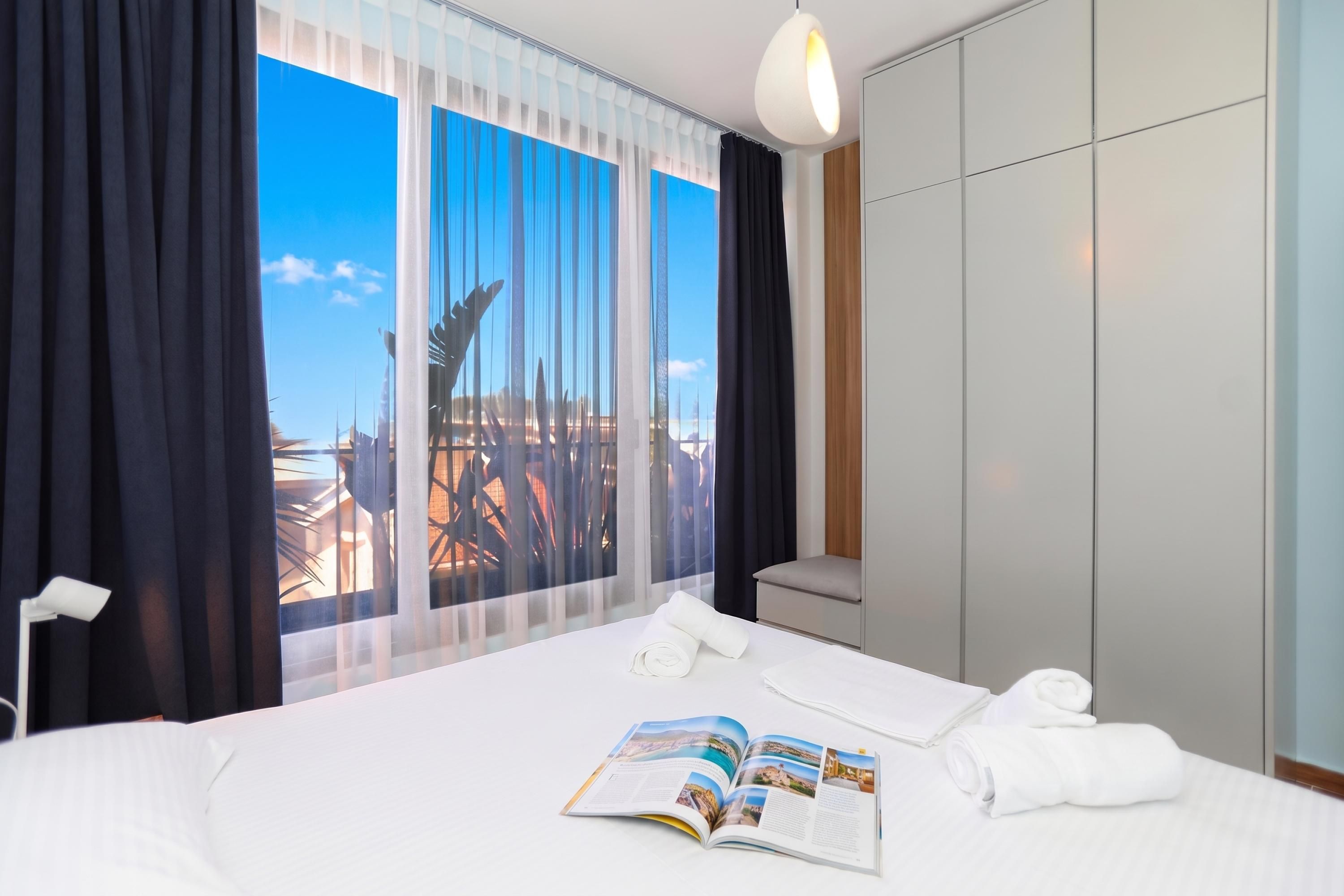 Een hotelkamer met een bed, een raam met gordijnen, een bedlamp en een tijdschrift op het bed.