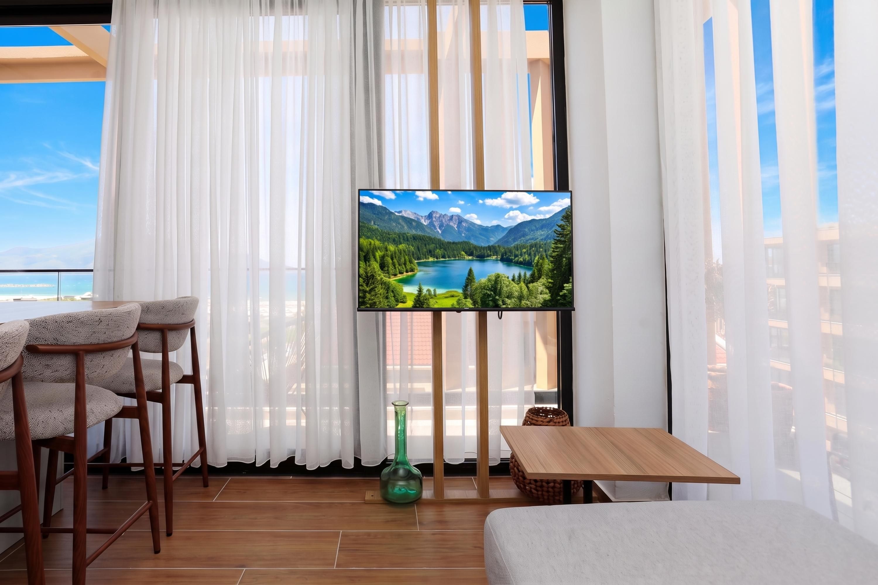 Una habitación con una televisión sobre un mueble bajo, una mesa de madera, dos sillas, un jarrón verde y cortinas de gasa.