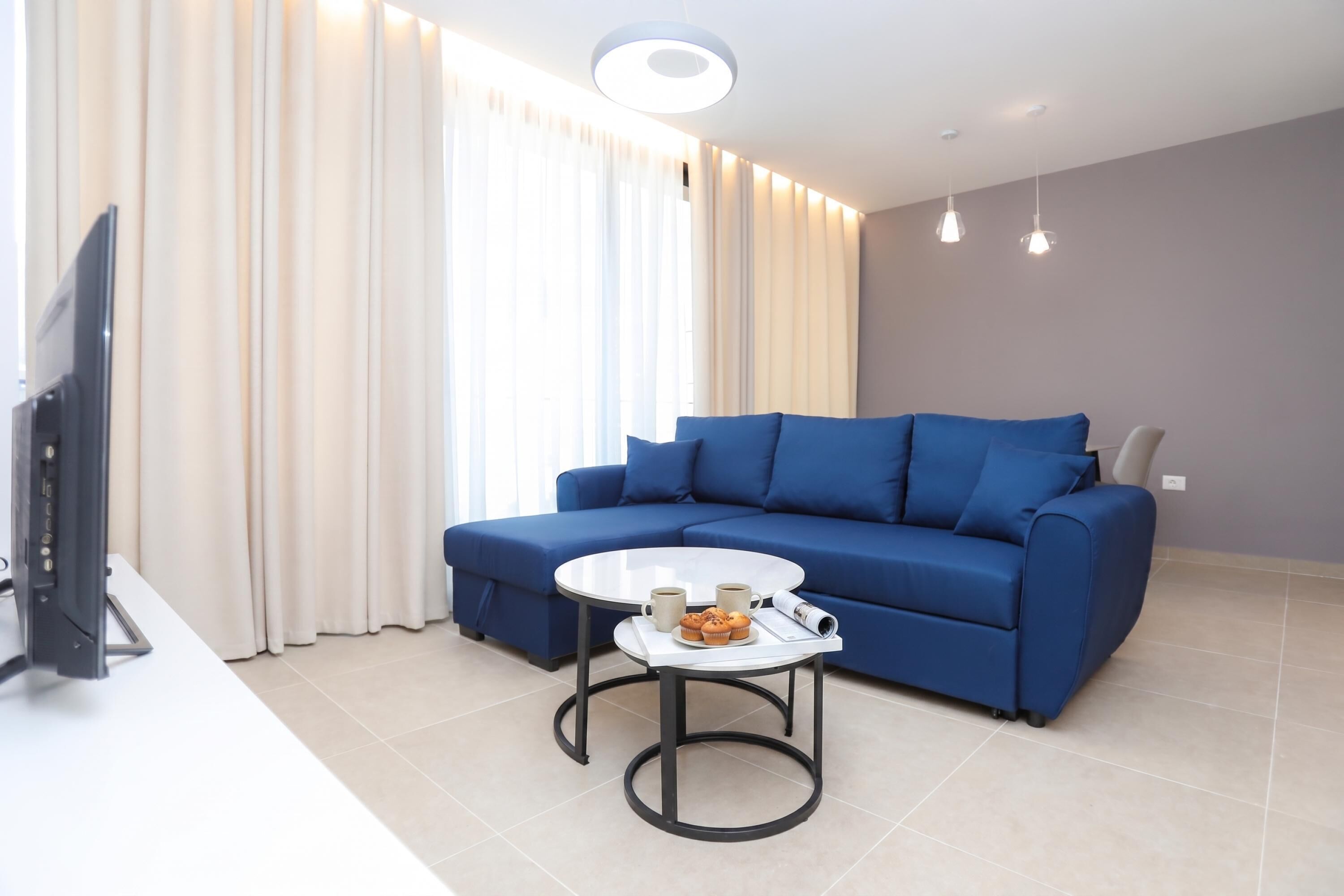 A modern living room with a blue sofa, a small round table with a coffee cup and muffin, and a flat-screen TV on a stand.