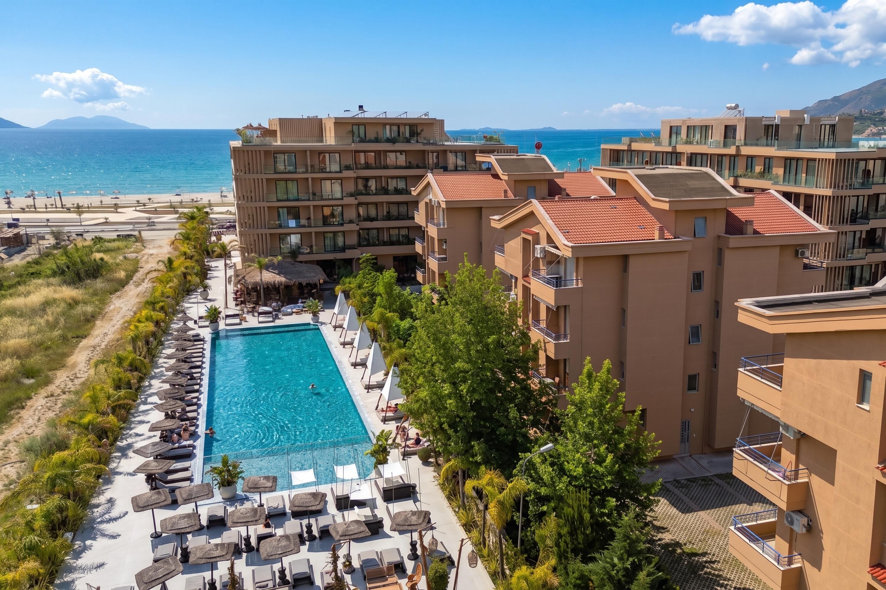 A hotel with a swimming pool surrounded by lounge chairs and umbrellas, overlooking a beach and the sea.