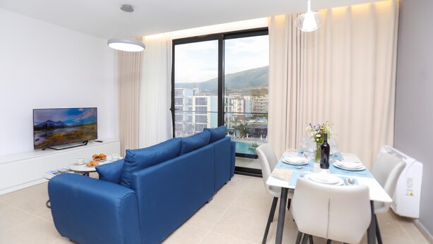 Un salon moderne avec un canapé bleu, une table à manger dressée pour deux personnes et une grande fenêtre donnant sur un paysage urbain.