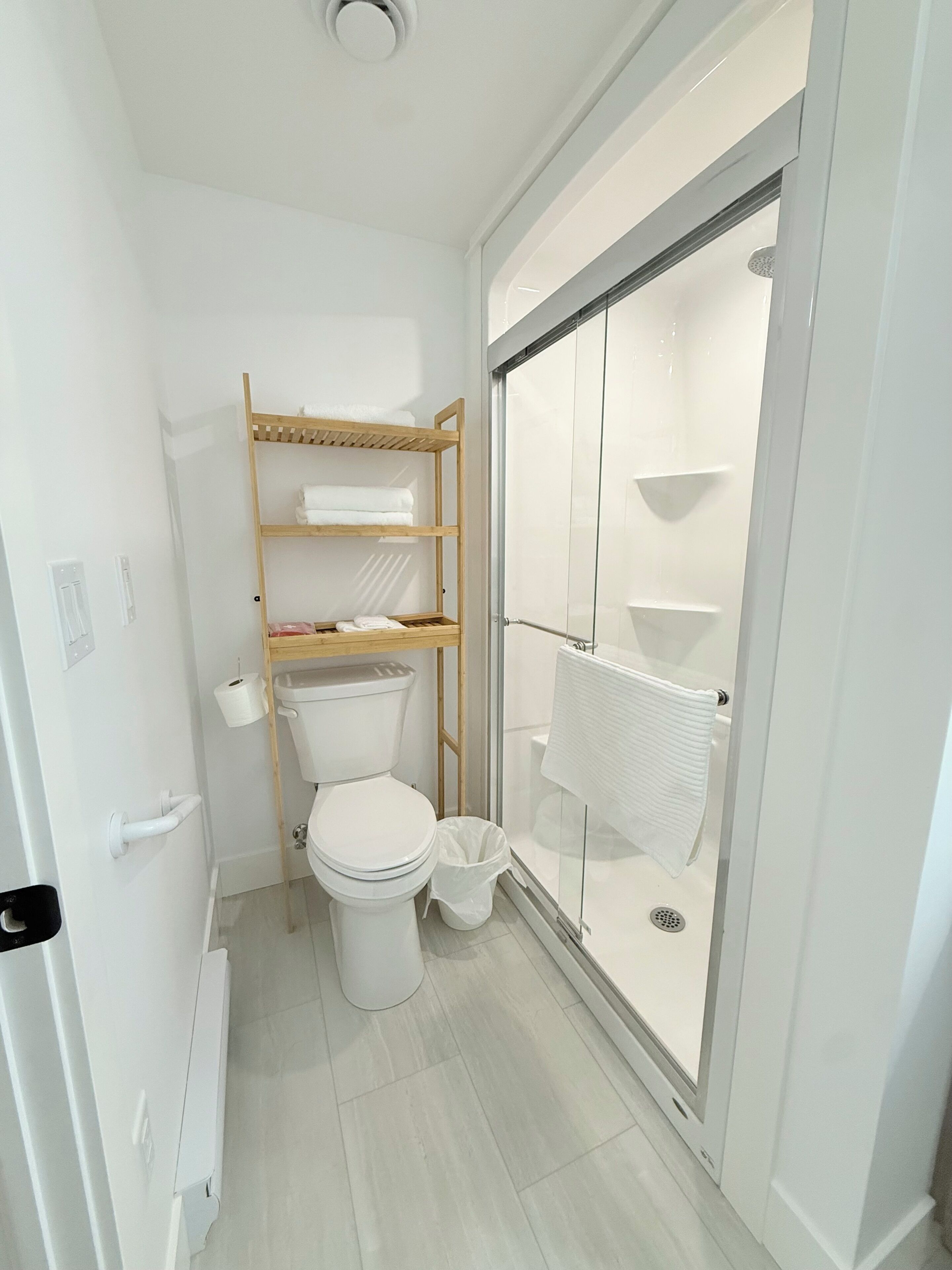 Une petite salle de bain comprenant des toilettes, une étagère en bois avec des serviettes et une cabine de douche en verre.
